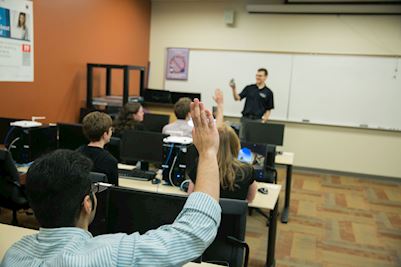 A student wearing a blue shirt raising his hand. We see him from behind. 